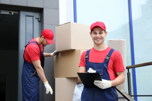 Two removal workers dressed in red caps and blue overalls are engaged in a house moving task outside a modern residential building. The worker on the left is focused on lifting or adjusting a large cardboard box, which is stacked on top of other similar boxes near the entrance. This individual is wearing white gloves and has their head tilted down towards the boxes, suggesting careful handling. The worker on the right is smiling directly at the camera, holding a clipboard and dressed similarly, with a supportive stance. The background includes a dark grey door and a section of the exterior wall, with large glass windows reflecting the surroundings. The setting appears to be on a residential street in Wimbledon, indicated by the subtly inferred location from the website context. The image captures the professional atmosphere of a house removals service, emphasizing careful handling of household items during a move, with a focus on teamwork and efficiency on a typical outdoor entryway scene.