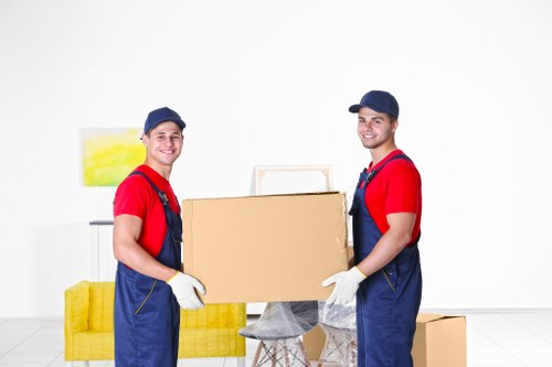 Two removal technicians from Wimbledon Man with Van, dressed in matching red shirts, blue overalls, and navy caps, are standing indoors against a plain white wall, engaged in a house moving task. They are holding a large cardboard box together, with both wearing white gloves. Behind them, a yellow upholstered sofa with a bright, abstract yellow and green painting hanging above it is visible on the left side of the image. To the right, there are additional cardboard boxes stacked on the floor, indicating an ongoing packing or moving process within a residential setting. The floor appears to be light-colored wood or laminate, highlighting a clean and organized environment. The lighting is bright and evenly distributed, emphasizing the professional and neutral tone of the scene. The overall context suggests a typical interior space being prepared for house removals or relocation services, illustrating the approach of Wimbledon Man with Van in serving residential moves in the Wimbledon area, possibly within SW19 postcode or nearby South West London suburbs.