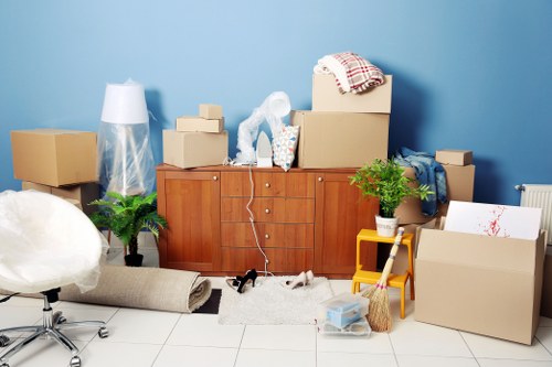 A collection of household items and moving supplies arranged against a light blue wall in a room, likely during a house move in Wimbledon. In the foreground, there are several cardboard boxes of various sizes, some sealed and others open, with packing paper and bubble wrap visible on top. A wooden sideboard or cabinet is positioned centrally, with a white table lamp, a potted green plant, and a roll of packing paper placed on and around it. To the left, a small, white office chair with a metallic frame is partially visible, and behind it, a transparent plastic bag filled with items stands upright. On the floor, there is a rolled-up rug or carpet, a small white rug, and a pair of black shoes. To the right, a yellow plastic step stool supports a stack of small boxes, with a cardboard box leaning against the wall. Additional packing materials, including a broom and a plastic storage box, are scattered around. The scene depicts a typical moving process within a residential space in the Wimbledon area, illustrating the packing stage of a professional removals service by Wimbledon Man with Van.