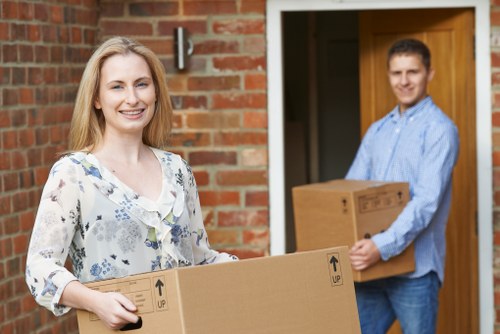 A smiling young woman with blonde hair and a floral blouse is standing outside a brick house, holding a medium-sized cardboard box with printed arrows indicating the correct orientation. In the background, a young man in a light blue shirt with rolled-up sleeves is standing near the doorway, holding a similar cardboard box. The scene captures the process of household relocation, with the exterior of a residential property and a visible wooden doorframe. The boxes appear to be filled with personal belongings and are made of brown corrugated cardboard. The lighting suggests natural daylight, and the setting reflects professional house removal services offered by Wimbledon Man with Van, based in the Wimbledon area, assisting clients with efficient and organized relocations within the local region.