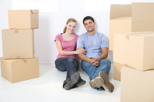 A young couple with light skin and casual clothing, sitting on the floor of a bright, minimalist room surrounded by moving boxes of various sizes made from brown cardboard, some stacked on top of each other. The woman is wearing a pink T-shirt and blue jeans, with blonde hair tied back, and the man is wearing a light blue T-shirt and dark jeans, with dark hair. They are smiling and holding hands, with the woman resting her arm on the man's shoulder. The boxes are placed around them, indicating a house removals or moving process typically seen in residential environments in Wimbledon or nearby areas. The room has a plain white wall and natural lighting illuminating the scene, emphasizing the theme of packing, moving, and relocation services that Wimbledon Man with Van provides for local households.