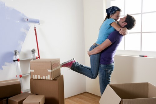 A man and woman engaged in a house removal within a bright, minimally furnished room, likely part of a residential property in Wimbledon. The woman, dressed in casual clothing with a blue top and jeans, is being lifted off the ground by the man, who is supporting her from beneath. The woman has her arms around the man's shoulders, and both are smiling and appear happy. The room features a large window allowing natural light to fill the space, with white walls and minimal decor. In the foreground, several cardboard boxes of varying sizes, some sealed and others open, are stacked and scattered on the wooden floor, indicating packing activity. A roll of packing paper or bubble wrap is attached to a wall-mounted dispenser, and the scene reflects the process of preparing household belongings for a move, typical of house removals services in Wimbledon. The image captures the focus on personal belongings, including boxes and the lifting activity, with a clean, well-lit interior environment suited to residential relocations in the area.