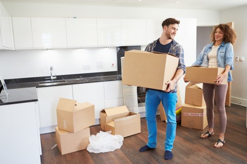 A man and a woman are inside a modern, well-lit kitchen with white high-gloss cabinets and a black countertop, engaging in a house move. The man, dressed in a casual plaid shirt and blue jeans, is carrying a large, plain cardboard box towards the right side of the image, smiling as he handles the item. The woman, wearing a light denim jacket and maroon trousers, stands nearby holding a smaller cardboard box, also involved in packing or unloading. On the dark wooden floor, there are multiple cardboard boxes of various sizes, some sealed and others open, with packing paper visible inside a few, indicating packing activity. The open-plan kitchen features a stainless steel sink, a contemporary faucet, and a dark backsplash extending behind the countertop. In the background, the room appears spacious with natural light enriching the neutral, modern aesthetic, representing a typical residential property around Wimbledon. The scene captures the process of packing or unpacking during a house removal, demonstrating professional movers from Wimbledon Man with Van handling household belongings efficiently, in a setting characteristic of homes within the SW19 postcode area.