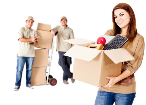 In the foreground, a young woman with long brown hair, dressed in a beige top and blue jeans, is smiling and holding a cardboard box filled with household items including a red apple, a black calculator, and some magazines, suggesting she is preparing for a house move near Wimbledon. Behind her, on a clean white background, two male movers dressed in beige work uniforms and caps are seen; one is holding a stack of packed cardboard boxes, while the other, using a hand truck or trolley, lifts a sealed box. The movers are positioned to the left, with additional boxes stacked nearby, indicating a professional removals service. The scene conveys an organized and efficient moving process, likely within a residential setting, with the focus on household items ready for transportation, reflecting the services provided by Wimbledon Man with Van in the Wimbledon area according to the webpage context on payment and security for house removals.