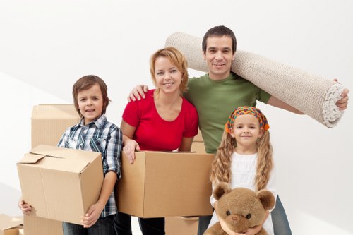 A family of four, consisting of a mother, father, a young boy, and a young girl, is posing together in a bright, plain indoor space. They are smiling and holding various household items and boxes, indicating they are in the process of moving or preparing for a house relocation. The mother, wearing a red top, has her arm around the young boy, who is dressed in a checkered shirt and holding a small cardboard box sealed with brown packing tape. The father, in a green T-shirt, stands next to the mother with a large rolled-up carpet resting on his shoulder. The young girl, in a white sweater and colorful headband, holds a teddy bear and stands to the right of her father. Behind the family, there are several cardboard boxes of different sizes, some stacked, and they all appear to be in a spacious, uncluttered household environment. The backdrop is plain white, emphasizing the subjects and the household items, which reflect typical elements involved in house removals, with a subtle nod to local residential areas such as Wimbledon or nearby postcode regions. The scene conveys a sense of preparation and teamwork associated with moving homes, with natural lighting highlighting their happy expressions.