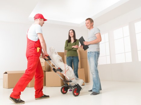 Interior of a bright, modern room with white walls and large windows, showing a professional removals team from Wimbledon Man with Van. The team member on the left, dressed in red overalls and a red cap, is pushing a hand truck loaded with wrapped household items and cardboard boxes. A woman standing in the middle, wearing casual green sweater and jeans, is holding a clipboard and smiling, while a man on the right, dressed in a light grey t-shirt and jeans, is engaged in conversation and holding a dark folder or notebook. Several large cardboard boxes are arranged around them, some stacked, indicating an ongoing house removal or packing activity. The scene captures the preparation phase of relocating household belongings within or from a property in Wimbledon, with natural daylight illuminating the space, highlighting the professional and organized approach of the furniture and item handling involved in the removals process.