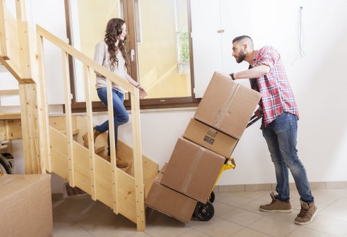A male removal worker wearing a red and white checkered shirt, jeans, and brown shoes is pushing a hand trolley loaded with three large, sealed cardboard boxes up a slight incline on a tiled floor inside a residential property. The boxes are stacked neatly, with the largest at the bottom and smaller ones on top. He is positioned near a wooden staircase with a natural finish, which has a simple handrail, leading to an upper floor. A young woman with long, curly brown hair, dressed in a white top and blue jeans, is standing at the base of the staircase, holding onto the handrail and watching the worker. Behind her, there is a closed glass door with a wooden frame that allows natural light to illuminate the room, which has a clean, neutral-toned interior. The environment suggests an ongoing house move or rearrangement, with the focus on safe handling and transport of household items, emphasizing a professional removals service typical of Wimbledon in south-west London.