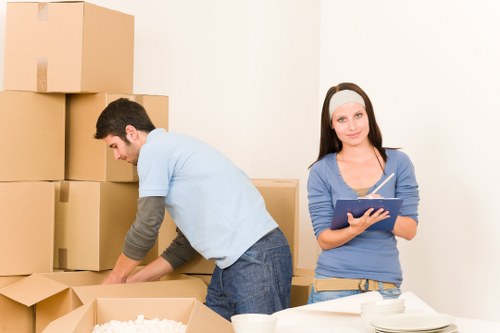 A young man with dark hair, dressed in a light blue t-shirt and jeans, is seen unpacking cardboard boxes in a residential interior, possibly a living room or hallway, with several stacked moving boxes around him. The boxes are made of brown corrugated cardboard, some sealed with packing tape, and contain household items and packing materials. To the right, a young woman with long dark hair, wearing a blue long-sleeve top, a pink headband, and light-colored pants, holds a clipboard and pen, standing near a white table that has a stack of white plates or bowls. The background features a plain white wall, with soft lighting suggesting an indoor environment typical of a house or apartment in Wimbledon, London. The scene reflects a house removal process managed by Wimbledon Man with Van, with the focus on household packing and organizing in preparation for a move within the local area.