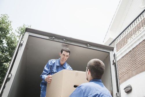 A professional movers wearing blue uniforms are unloading a large cardboard box from a grey removals van parked outside a residential property. The interior of the van is visible, showing the open back door and spacious cargo area. One mover, standing inside the van, is smiling and holding the box, while another, with only the back of his head visible, is assisting from outside. In the background, the external environment includes part of a brick house with a white balcony railing and a residential street setting, typical of Wimbledon or nearby areas. The scene is well-lit with natural daylight, emphasizing the careful handling of household items such as the cardboard box, likely filled with personal belongings or household goods for a house move. This image subtly reflects the professional house removals services offered by Wimbledon Man with Van, highlighting their focus on safety and efficiency during the relocation process in London or its surrounding postcodes.