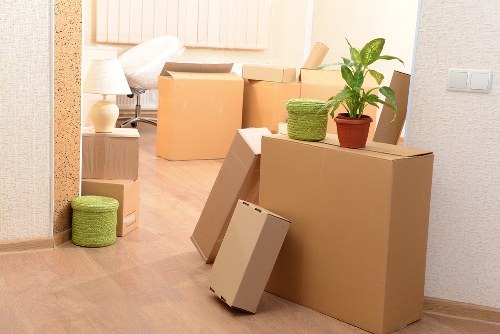 The image shows a residential interior with a hallway or living area in the process of a house move, associated with Wimbledon Man with Van's removals services. Several cardboard boxes of various sizes, some sealed with packing tape, are arranged on the wooden floor in front of a wall with a textured wallpaper. A large flat cardboard box is leaning against a smaller one, while another box is placed upright nearby. On top of a sizeable cardboard box, there is a small potted plant with green foliage and a decorative woven basket or container, suggesting the storage of household items or personal belongings. To the left, a green cylindrical storage ottoman made of woven material is positioned on the floor. The background reveals a glimpse into a bedroom or another living space, with a bed frame and a bedside table holding a lamp, indicating the area belongs to a private home in Wimbledon or nearby. The scene is well-lit, emphasizing the natural tones of the wooden flooring and neutral-colored walls, embodying typical property contents associated with local house removals and sustainable moving practices.
