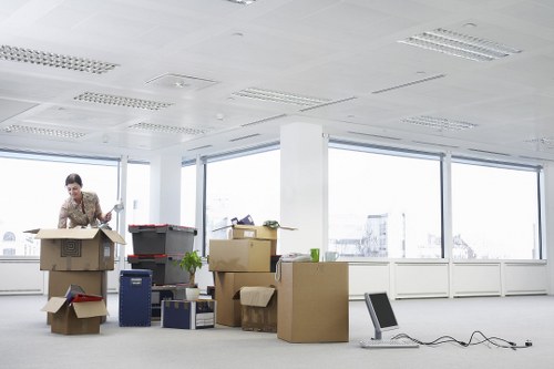 A woman in a patterned blouse is standing in a spacious office with large floor-to-ceiling windows that reveal a bright, overcast sky outside. She is surrounded by numerous cardboard boxes, some open with packing paper visible, and others sealed with packing tape, indicating recent or ongoing packing activities. The boxes are of varying sizes and are positioned on the floor, with some stacked or leaning against each other. Various office items are scattered around, including a potted plant, a blue plastic storage container, and a small cardboard drawer unit. An office chair is placed at a slight distance, accompanied by loose cables draped across the floor. The interior features a white ceiling with recessed lighting, and the room’s walls are primarily glass, offering an unobstructed view outside, suggesting a commercial environment in Wimbledon or nearby. The scene depicts a preliminary stage of house or office removals, fitting within professional relocation or removals services provided by Wimbledon Man with Van.