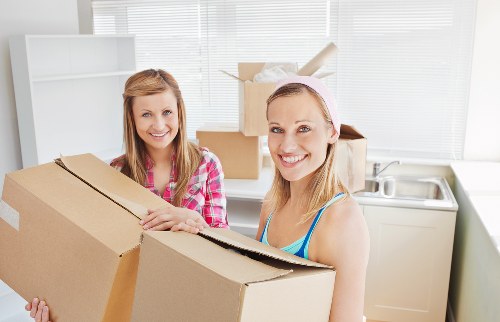 Two women with light skin and blonde hair are indoors, smiling at the camera while holding a large, partially opened cardboard box during a house removal in Wimbledon. One woman, dressed in a pink and white checkered shirt, has her hands on the box, while the other, wearing a pink headband and a light blue sleeveless top, holds the box from the side. Behind them, a white kitchen unit with a sink and a window with closed blinds allow natural light into the space. Several other cardboard boxes, some closed and others open, are stacked or placed on the kitchen counter. The background shows a narrow hallway and the edge of a staircase leading to an upper floor. The scene portrays a professional house removal activity, with visible household items such as cardboard boxes and packing materials, reflecting a typical residential environment associated with local removals services in Wimbledon. Wimbledon Man with Van is experienced in handling such household items, ensuring safe packing and transport during relocations in the SW19 postcode area.