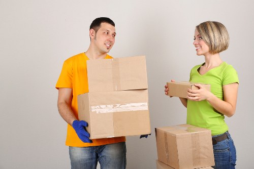 A male removal worker wearing a bright yellow shirt and blue gloves is holding two cardboard boxes stacked one on top of the other, while a female colleague dressed in a lime green top and jeans is smiling and holding a smaller cardboard box. They are standing indoors against a plain light grey wall, with additional cardboard boxes placed on a surface nearby. The scene depicts a professional house removals service, with the items suggesting they are in the process of packing or unloading belongings. The environment appears to be inside a residential property, possibly a living room or hallway, with minimal background detail. The lighting is neutral and even, highlighting the details and textures of the boxes and clothing. This image exemplifies services offered by Wimbledon Man with Van, focusing on local house removals in London, specifically Wimbledon area, through a clear depiction of furniture and packed items in a professional setting.