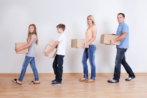 A family of four is inside a bright, modern house with light-colored walls and a wooden floor, engaged in packing or unpacking. The youngest girl in the front, wearing a grey top and jeans, carries a medium-sized cardboard box, while her older brother behind her, dressed in a white t-shirt and dark trousers, also holds a box. The mother, with blonde hair, is positioned further back, holding a medium-sized cardboard box, wearing a peach top and blue jeans. The father, wearing a light blue shirt and dark trousers, stands at the rear, carrying another cardboard box. The scene appears to be in a spacious hallway or living area, with no visible furniture or decorations, emphasizing the focus on moving activities. The family members are aligned in a line, spaced evenly, with the boxes varying in size but consistent with household relocation. The natural lighting highlights the neutral interior environment, supporting the context of house removals in Wimbledon or nearby areas, with a subtle emphasis on local residential surroundings and the professional service provided by Wimbledon Man with Van.