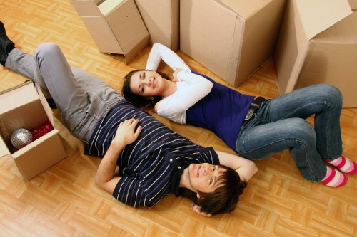 A man and a woman lie on a wooden floor surrounded by open cardboard moving boxes, indicating a house relocation in progress. The man, dressed in a striped dark blue and white shirt with grey trousers, is lying on his back with one arm resting on his stomach and the other behind his head. The woman, wearing a navy blue top with white sleeves and grey jeans, is lying on her side with her head resting on her hand, appearing relaxed. Several cardboard boxes around them vary in size, some containing household items such as a red and silver ball, while others are empty or partially packed, suggesting an ongoing decluttering or packing process. The background shows a residential interior with a plain wall and a part of a doorway, subtly indicating a dwelling within the Wimbledon area, consistent with professional removals services by Wimbledon Man with Van. The scene is lit with natural or ambient indoor lighting, creating a calm and neutral atmosphere typical of household moving situations in southwest London.