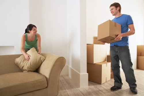 In a well-lit, modern interior space, a woman with dark hair tied back, wearing a green sleeveless top, is sitting on a beige sofa and holding a light-colored cushion, smiling towards a man who is standing and holding a medium-sized cardboard moving box. The man, dressed in a blue T-shirt and dark jeans, is smiling back and appears to be about to place or lift the box into a stack of similar cardboard boxes behind him, indicative of a household move. The background shows a collection of neatly stacked cardboard boxes of varying sizes, some partially visible, arranged against a white wall with a clean, minimalist design. The light wood flooring complements the neutral tone of the furniture and boxes, suggesting a typical residential setting in Wimbledon or nearby areas. The scene captures a moment of packing or unpacking, characteristic of house relocation services offered by Wimbledon Man with Van, with emphasis on household items and the process of moving home sustainably.