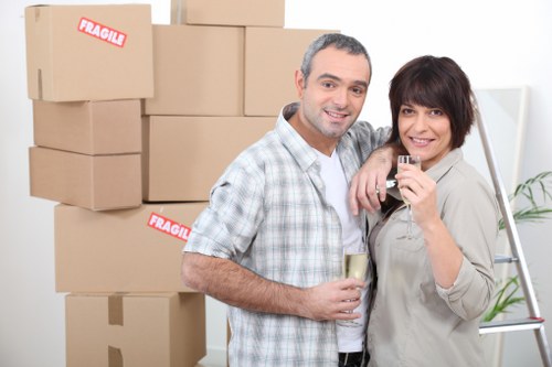 A middle-aged man and woman stand close together indoors, smiling at the camera, with the man holding a glass of white wine and the woman holding a small glass, possibly containing a beverage. Behind them is a stack of cardboard boxes, with some labeled 'Fragile,' indicating they are preparing for a house move. The boxes vary in size and are arranged to the left side of the image, in a well-lit environment with natural or bright artificial lighting. The man has short, dark hair with some grey, and is dressed in a checkered shirt with rolled-up sleeves. The woman has short, dark, layered hair with bangs and wears a light-colored shirt with rolled-up sleeves. To the right, a step ladder with a potted plant on the top step is visible, suggesting a domestic setting, likely a living room or hallway in a house or flat in Wimbledon. The scene reflects a positive, celebratory moment during a domestic relocation, supported subtly by the mention of house removals and local context.