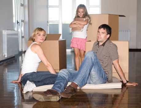The image depicts a family scene inside a well-lit home, with three individuals positioned on a polished wooden floor surrounded by open cardboard boxes, indicating a house move or unpacking process. A woman with blonde hair, wearing a white sleeveless top and blue jeans, is seated on the floor to the left, with her legs extended and her hands resting on the floor behind her. A young girl with long hair, dressed in a pink skirt and white top, stands behind a cardboard box, crossing her arms and gazing forward. To the right, a man with dark hair, wearing a striped shirt and jeans, is seated on the floor leaning back on his hands, with his legs stretched out and crossed at the ankles. The background shows a bright room with windows, radiators, and additional cardboard boxes stacked behind the family members, suggesting they are in the process of moving into or out of a property in Wimbledon or nearby. The scene emphasizes household furniture, personal belongings, and packaging materials typical of domestic relocation services provided by Wimbledon Man with Van in the Wimbledon area.