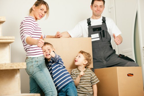 A family of four, including a woman and two young children, are inside a bright, modern home with white walls, standing next to a partially packed cardboard box. The woman, dressed in a striped long-sleeve shirt and jeans, is smiling and giving a thumbs-up gesture, while two children, a boy in a blue and white striped shirt and a girl in a beige and brown striped top, are standing close to her, with the boy giving a thumbs-up and the girl looking up. In the background, a professional house mover from Wimbledon Man with Van, wearing a white T-shirt and black overalls, is standing next to a large cardboard box, giving a thumbs-up as well. The scene appears to be part of a house removal or packing process, with the environment well-lit by natural or overhead light, and the setting within a residential property that suggests a typical London suburb, potentially near Wimbledon or the SW19 postcode area. The items and people are positioned on a staircase landing or open-plan hallway, emphasizing an active, organized moving scenario.