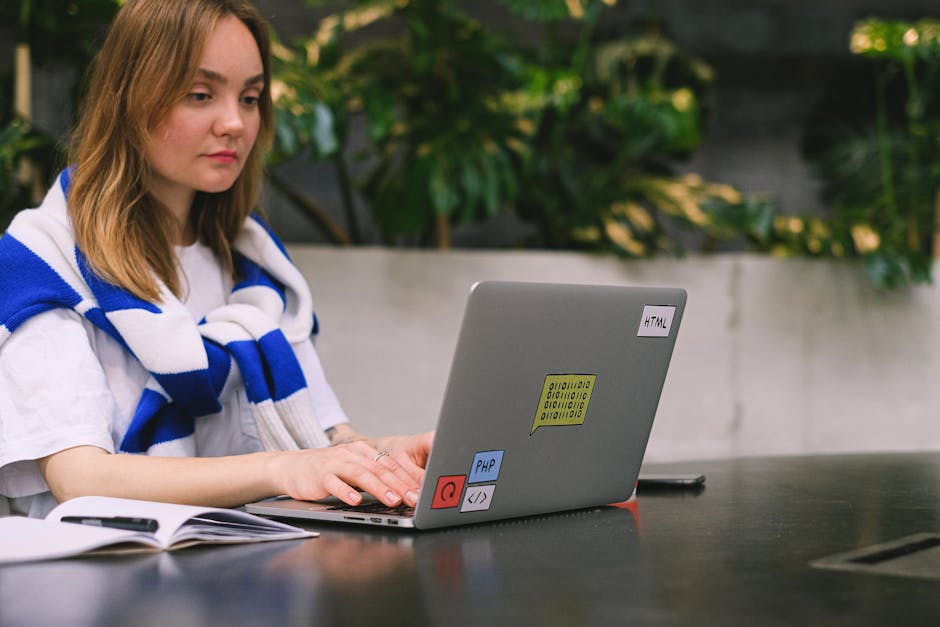 A young woman with shoulder-length light brown hair and a neutral expression is seated at a black table in a well-lit indoor space, with a backdrop of green leafy plants and a white wall. She is weari