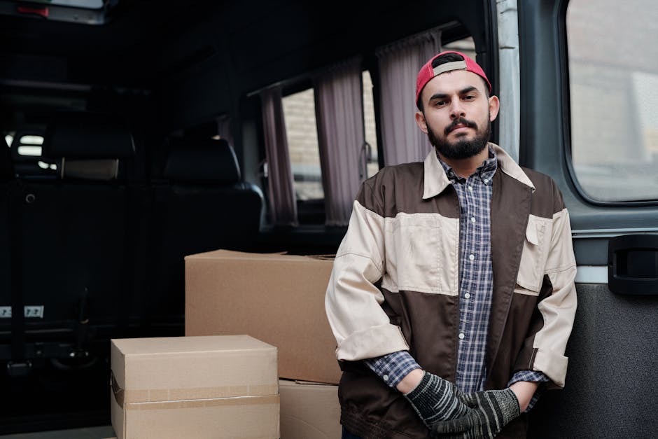 A man sitting on the ground in front of an open van, which is loaded with neatly stacked cardboard boxes of various sizes, ready for a home relocation. The man is wearing a blue beanie, a navy and red
