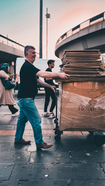 A man wearing a black long-sleeved shirt and blue jeans is seen loading a large wooden cart filled with stacked flattened cardboard boxes and wrapped items onto a trolley. The scene takes place outdoo