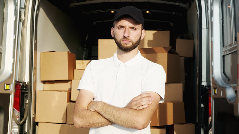A man wearing a black long-sleeved shirt and blue jeans is seen loading a large wooden cart filled with stacked flattened cardboard boxes and wrapped items onto a trolley. The scene takes place outdoo